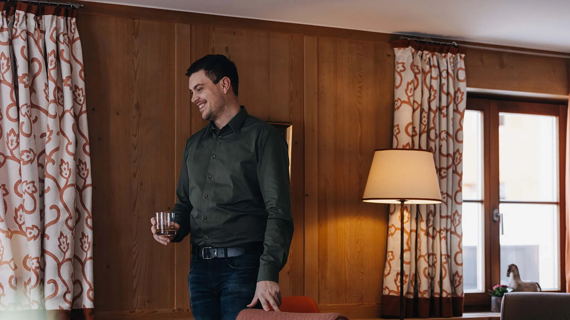 Mann mit Getränk steht lächelnd in gemütlichem Wohnzimmer mit Holzwänden