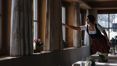 Frau in Tracht öffnet Gardinen in einem gemütlichen Gastraum