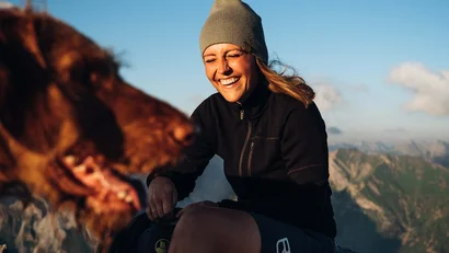 Frau mit Mütze lacht mit braunem Hund in den Bergen bei Sonnenschein