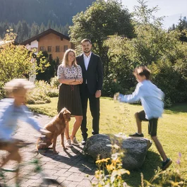Familie im Garten mit Hund, zwei spielende Kinder, Haus und Berg im Hintergrund
