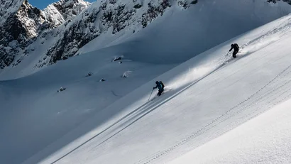 Zwei Skifahrer fahren einen verschneiten Berghang mit Bergen im Hintergrund hinab