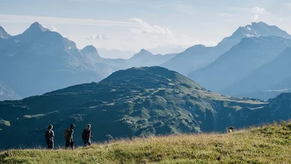 Drei Wanderer auf einem grasbewachsenen Hügel mit Bergen im Hintergrund