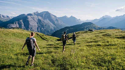Drei Wanderer auf einem grünen Bergpfad mit Bergpanorama im Hintergrund