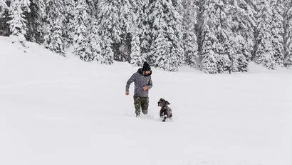 Mann läuft mit Hund im tiefen Schnee vor schneebedecktem Wald