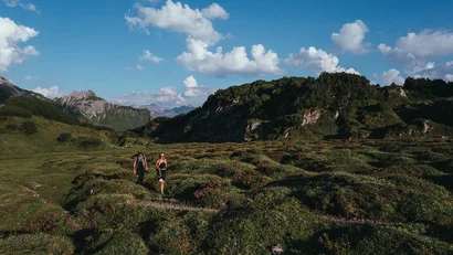 Zwei Wanderer auf Bergpfad in grünem alpinem Gelände unter blauem Himmel