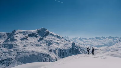 Zwei Personen stehen auf schneebedecktem Berggipfel mit Alpenpanorama