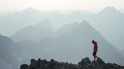 Mann in roter Jacke auf felsigem Berggipfel mit nebeligen Bergen im Hintergrund