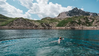 Person schwimmt in klarem Bergsee mit Berglandschaft und blauem Himmel